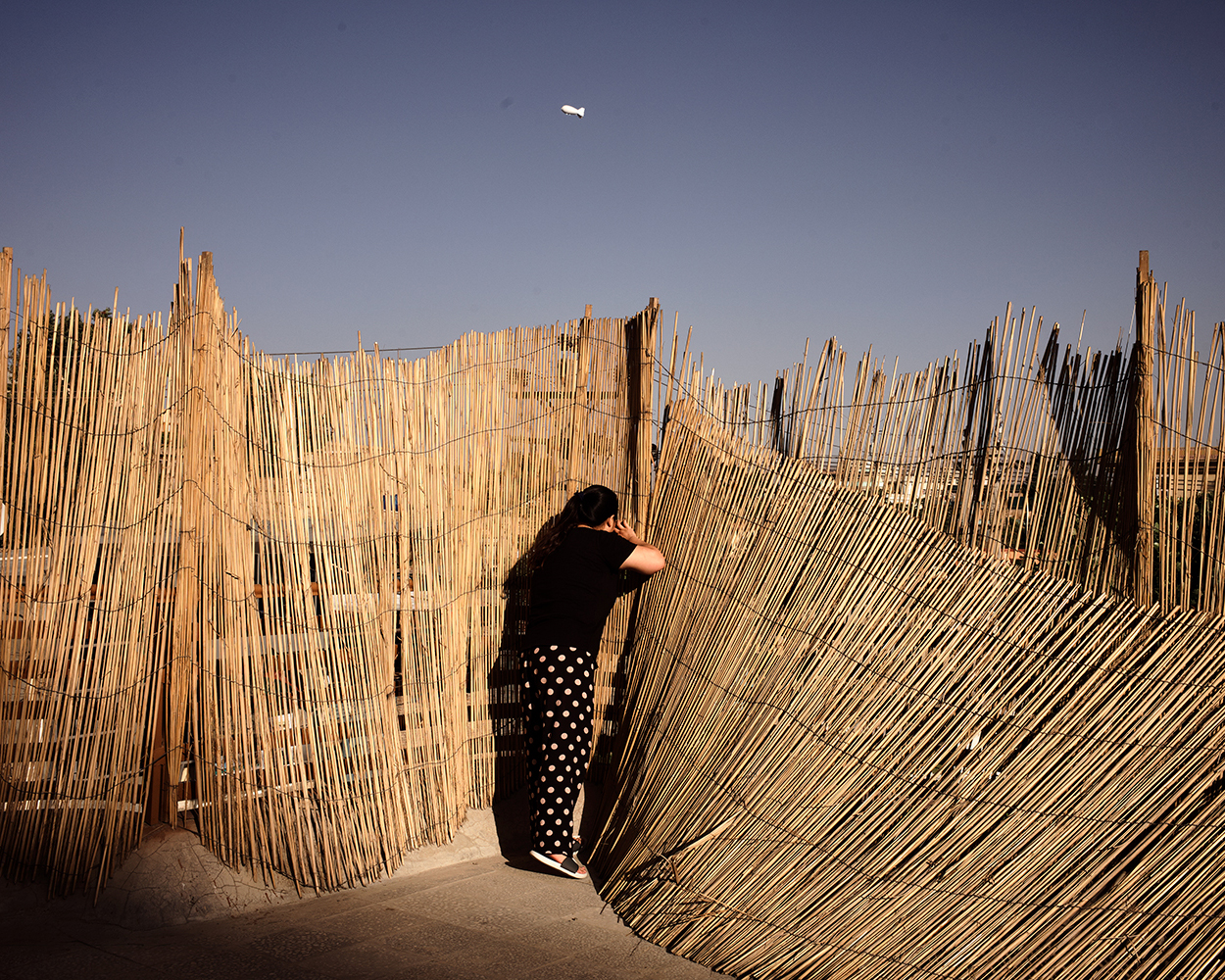 2018年，喀布尔中国饭店的天台上，红姐透过木栅的缝隙看外面。富人区的每家都很谨慎，布置起一些障碍物不让外界直视内部。天空中白色的点是一个汽艇，用来监视全城。