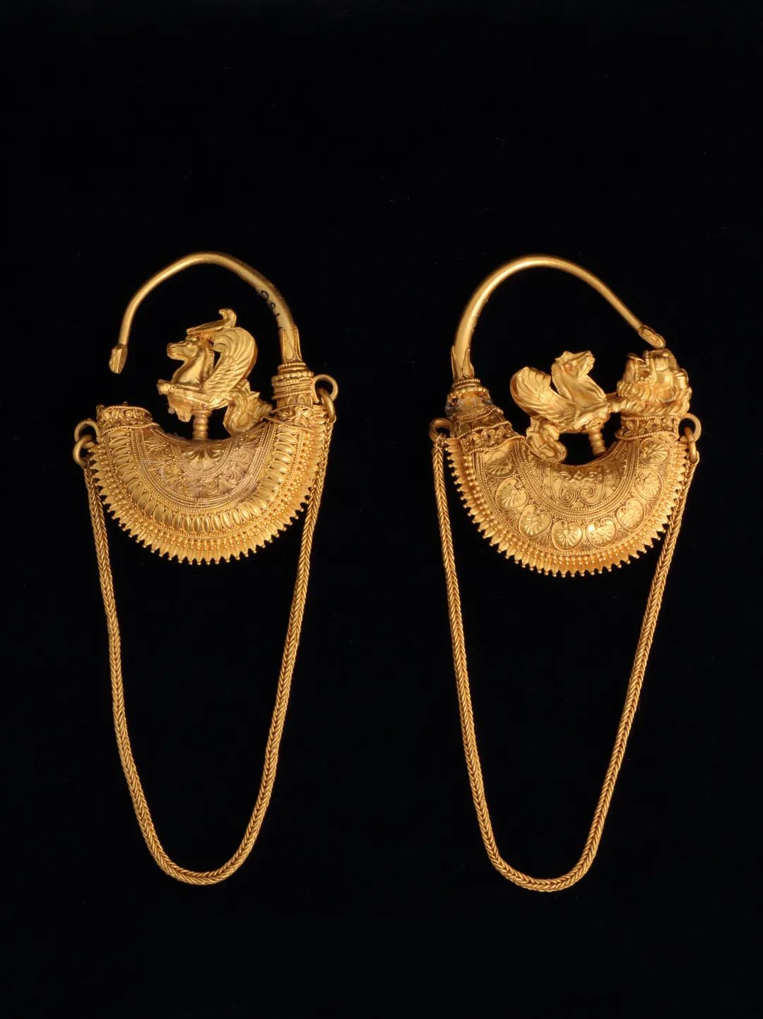 耳环一对（虚拟展出），约公元前375-前325年，黄金，霍莫莱之城（古马格尼西亚）出土，马格尼西亚古物局/沃洛斯考古博物馆藏（M 49， M 50）；图片来源：上海博物馆 耳环呈船型，尾端饰有金丝细工的狮首，船身上栖息有翼海马和小鸟。耳环以前后两半构成，上下端接合处均覆盖一排排细密的金珠颗粒。海马由前后两片金箔制成，经过切割并以浅浮雕装饰，狮首则是单独制作的。长金链松散地垂下，两端由靠近船顶部的圆环固定