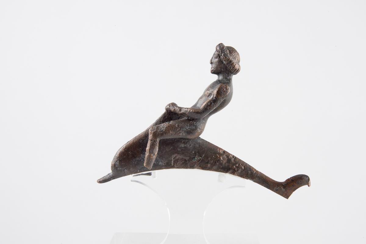 《骑海豚的青年》（非现场展品），公元前 475-450 年，青铜，雅典卫城出土，雅典卫城博物馆藏（EAM X 6626）；展览以全息投影呈现。 该像1888年发现于帕特农神庙东南部，是一件被献给雅典卫城圣殿的青铜容器上的装饰物。一位赤身裸体的青年正骑在一只海豚上，左手抓住它的背鳍。年轻男子与这只鲸目动物间的反向动作表现出他们在海上航行时的自然主义和激烈感。这一主题在古希腊艺术中尤为流行，因为它与海豚从海上冒险中拯救人类的神话有关。